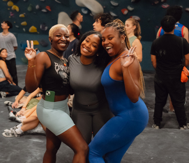 Three women pose smiling and flashing peace signs at Brooklyn Climbing Yoga and Fitness, surrounded by other people. They wear athletic outfits and appear happy and energetic, with climbing holds visible on the wall in the background.