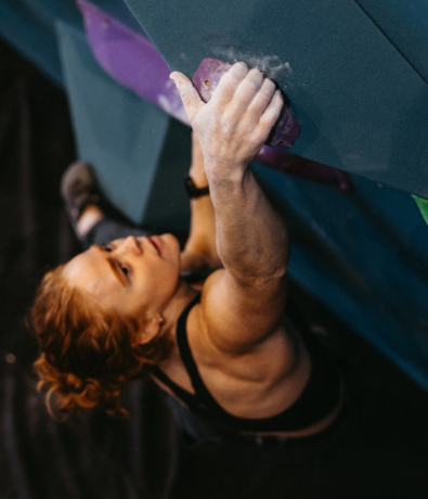 A woman with chalky hands and red hair climbs an indoor bouldering wall at Brooklyn Climbing Yoga and Fitness, gripping a purple hold while looking up with focus and determination.