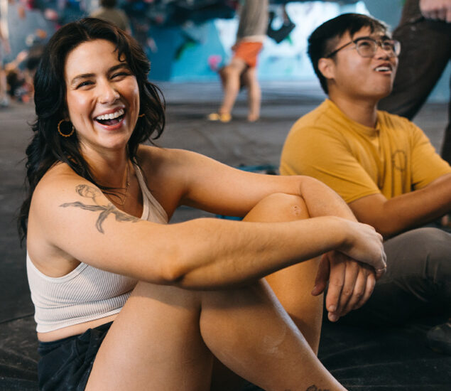 A woman in a white tank top and shorts sits on the floor smiling, with a man in a yellow shirt behind her. They are in an indoor rock climbing gym, with climbing walls and people in the background.