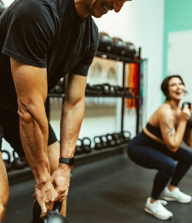A man and a woman exercise in a gym, each holding a kettlebell and performing squats. The man is in the foreground, smiling, while the woman in the background is also smiling. Gym equipment is visible behind them.
