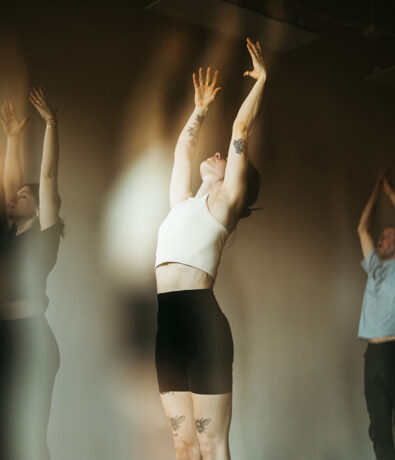 Several people practice yoga indoors, reaching their arms upward. The focus is on a woman in a white tank top and black shorts with tattoos on her arms and legs. Warm light and soft shadows create a calm atmosphere.