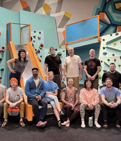 A group of people from local Community Clubs pose and smile together in front of an indoor climbing wall in Minneapolis, surrounded by colorful holds, geometric patterns, and a playful slide. Some are seated while others stand proudly.