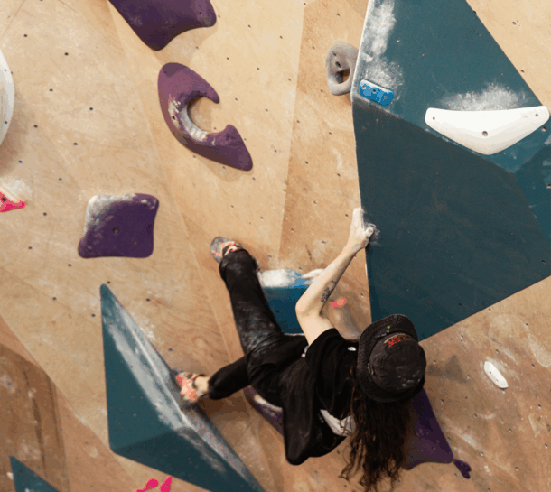 A person wearing a black shirt and cap is climbing an indoor bouldering wall in the Design District, gripping large blue and purple holds with their hands and feet. Chalk marks are visible on the wall and holds.