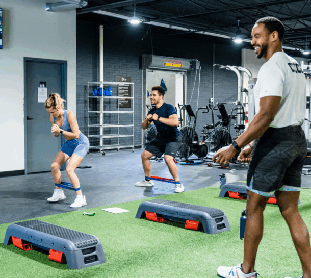 Three people exercise in a Design District gym using resistance bands around their legs, performing squats and lunges while smiling. Step platforms and gym equipment are visible in the background.