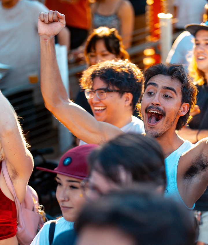 A diverse crowd of people outdoors at Boulderfest 2024 in Austin, smiling and cheering excitedly, with some raising their arms in celebration. The sunlight adds a warm, lively atmosphere to the scene.
