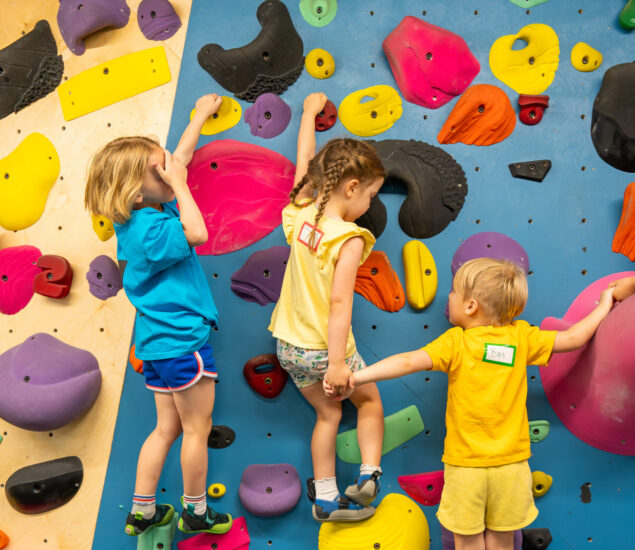 Three young children in colorful clothes climb an indoor rock wall with bright holds—a playful, energetic scene that highlights youth activities and teamwork, perfect for youth programs in DC.