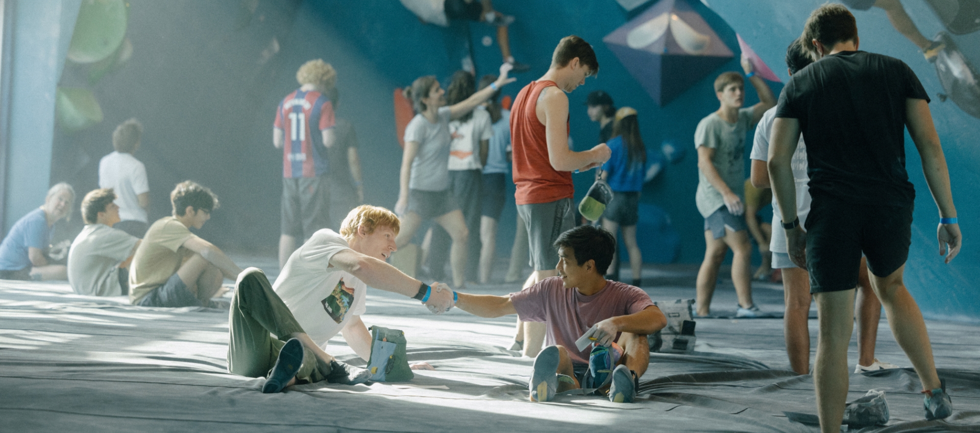 A group of people relax and interact on mats in an indoor rock climbing gym, with two young men in the foreground smiling and bumping fists while holding chalk bags. Others chat or prepare to climb in the background.