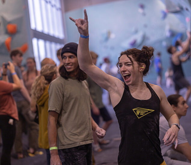A young woman in a black tank top smiles and raises her hand in a celebratory gesture at an indoor climbing gym during a Climb for Community event, surrounded by other climbers and supportive onlookers.