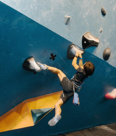 A young boy climbs an indoor bouldering wall at a Seattle climbing gym, gripping large black holds with arms and legs stretched out, wearing a harness, chalk bag, and climbing shoes—perfect for after school clubs.
