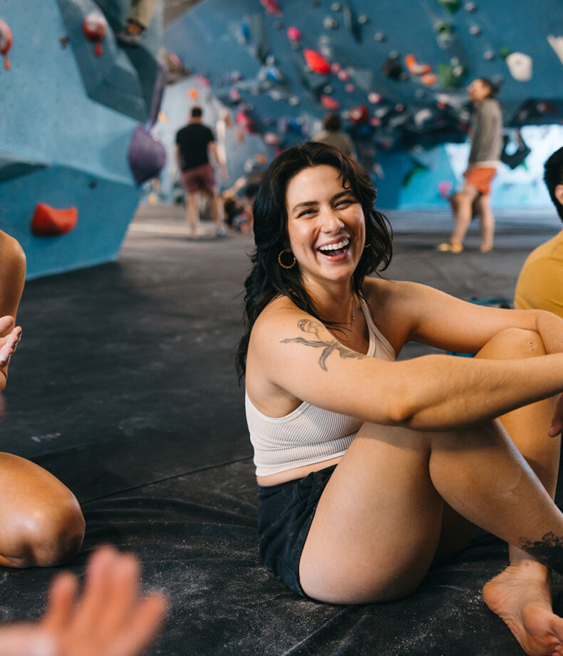A smiling woman sits on the floor of Climbing Tempe’s indoor gym, arms resting on her knees. Nearby, a man in a yellow shirt relaxes as colorful climbing holds fill the background.