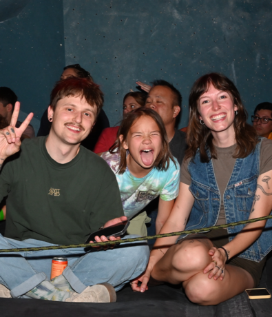 Three people sit and smile at the camera during Community & Events at Austin Bouldering Project; the man on the left flashes a peace sign, a child in the center makes a silly face, and the woman on the right smiles. Others relax in the background.