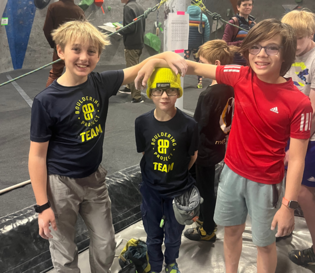 Three boys stand together at an indoor climbing gym. Two smile with arms around a third, shorter boy, who is wearing glasses, a hat, and a Bouldering Project Team shirt—part of the vibrant Boston youth teams community. Other kids are in the background.