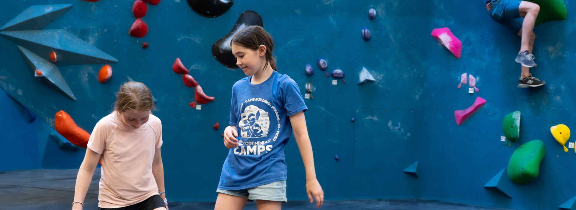 Two girls stand in front of a blue indoor climbing wall in Boston while a child climbs in the background. The colorful holds and geometric shapes set the scene as the girls discuss their plans, possibly preparing for Youth Teams practice.