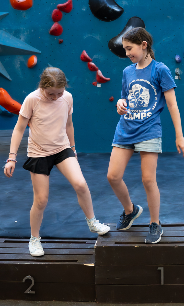 Two girls from Youth Teams stand on wooden platforms numbered 1 and 2 in front of a blue climbing wall with red holds. One wears a pink shirt and black shorts, the other a blue shirt and denim shorts—both representing Boston spirit.