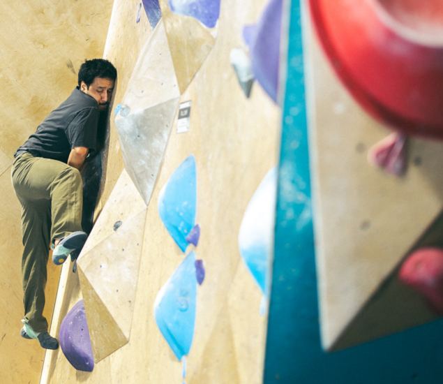 A man in olive pants and a dark shirt climbs an indoor bouldering wall at the 2025 DC Slab Showdown, gripping purple and blue holds while balancing on an angled, geometric surface.