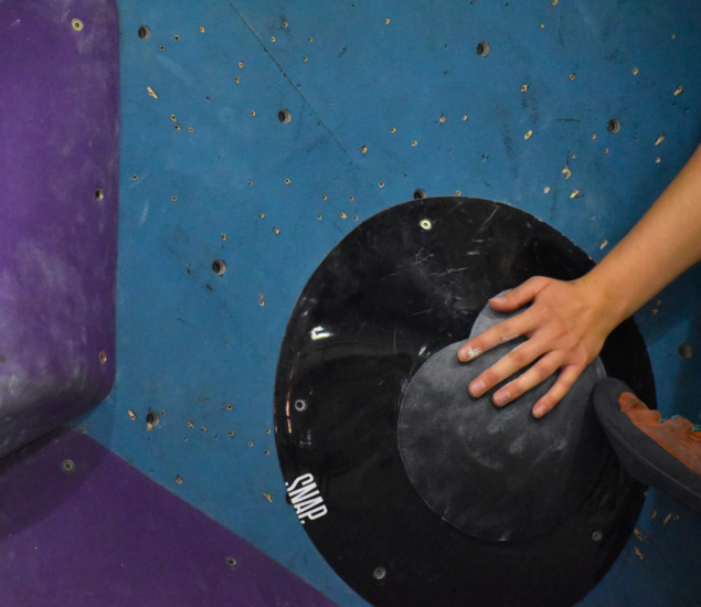 A person’s hand and climbing shoe grip a large, round black climbing hold on a blue indoor bouldering wall at the DC Slab Showdown 2025, with a purple hold visible to the left.