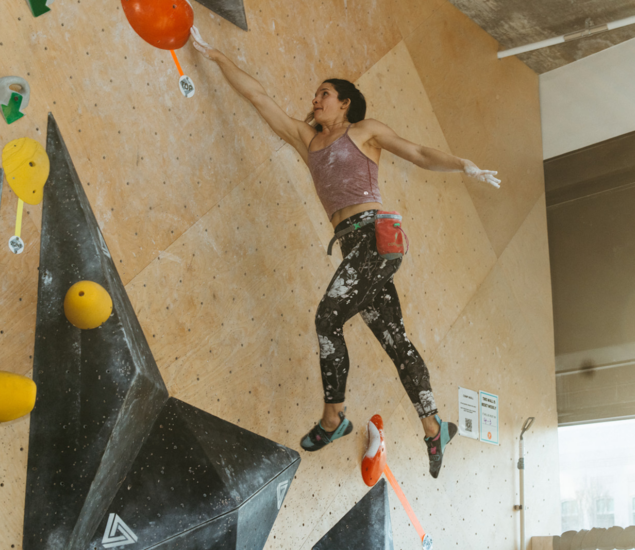 A woman in athletic clothing climbs an indoor bouldering wall during the 2025 Slab Showdown in DC, reaching for a large red hold with chalk-dusted hands and pants, her chalk bag at her waist.