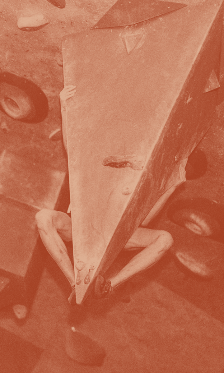 A rock climber, partially hidden behind a large, triangular climbing hold on an indoor wall, grips tightly—only legs and hands visible.