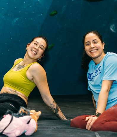 Two women enjoying their first time visit to the climbing gym, smiling on the floor in front of a blue wall with green holds. One wears a yellow top, the other a light blue shirt. Climbing gear and shoes are scattered nearby.