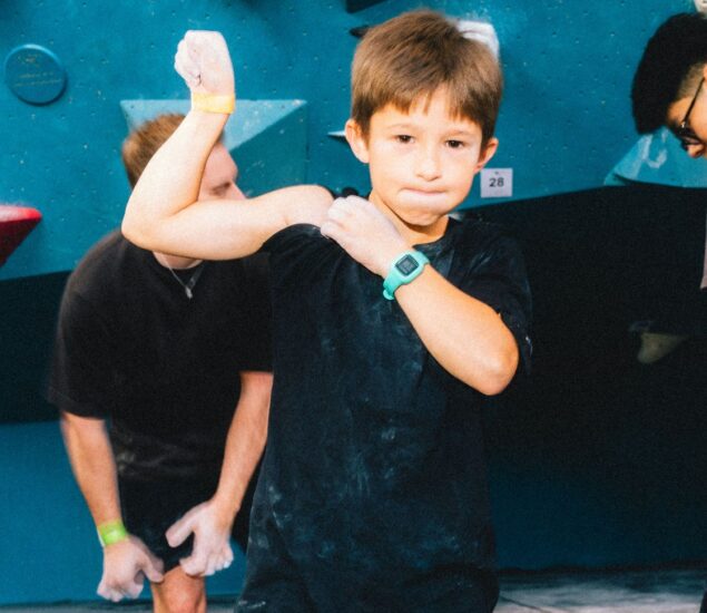 A young boy in a black shirt stands confidently in front of a climbing wall during Parents Night Out, flexing his arm and showing off his green wristwatch. Two people are in the background preparing to climb.