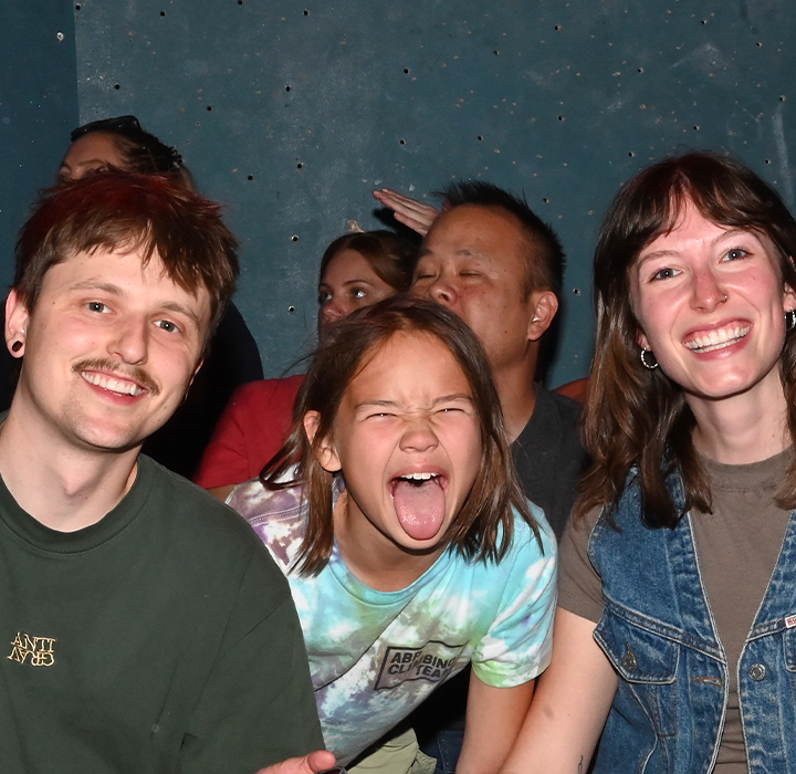 A group of people sits closely together in a crowded indoor setting, enjoying Membership Benefits at Austin Bouldering Project. In the foreground, a man gives a peace sign, a child sticks out their tongue playfully, and a woman smiles widely.