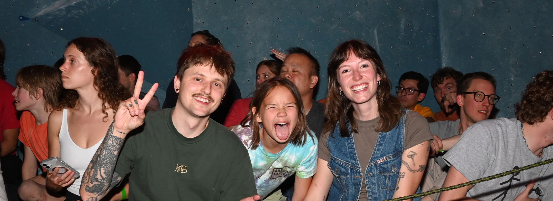 A group of people sits closely together in a crowded indoor setting, enjoying Membership Benefits at Austin Bouldering Project. In the foreground, a man gives a peace sign, a child sticks out their tongue playfully, and a woman smiles widely.