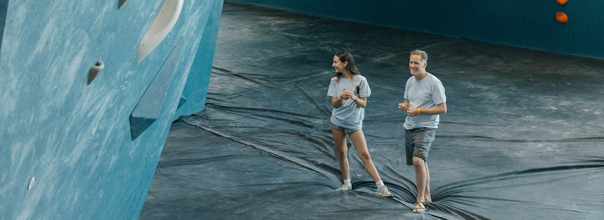 Two people in casual clothes stand on padded flooring at Austin Bouldering Project, smiling as they look up at a blue climbing wall. They appear relaxed, ready to enjoy the membership benefits at Austin Bouldering Project.