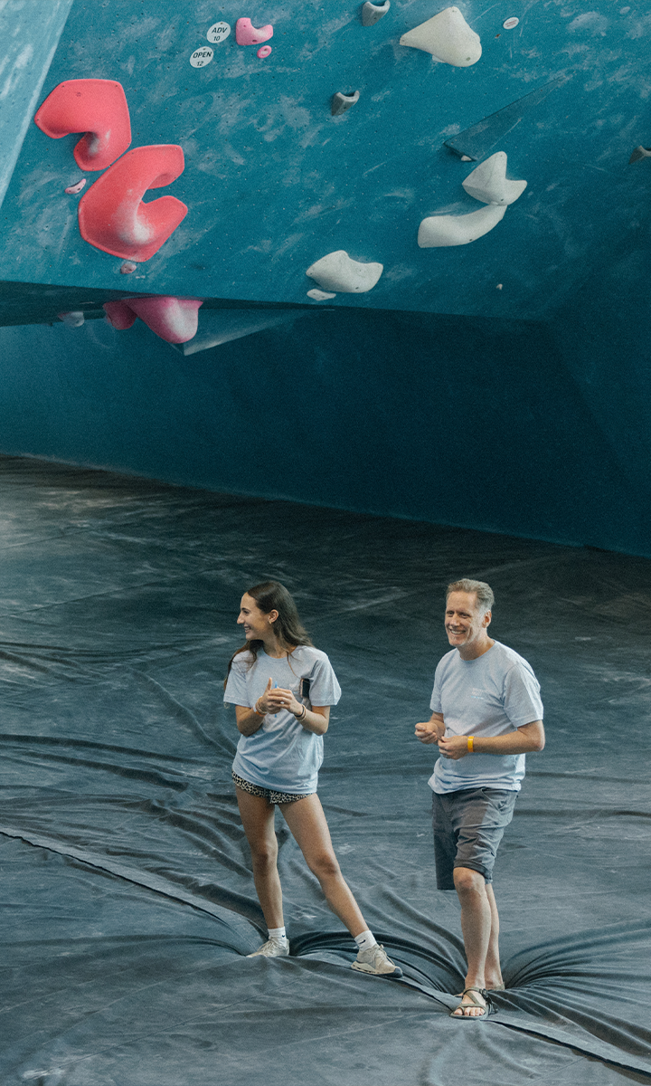 Two people in casual clothes stand on a padded floor, smiling and looking to the side, in front of a large indoor climbing wall with colorful holds—enjoying the Membership Benefits at Austin Bouldering Project.