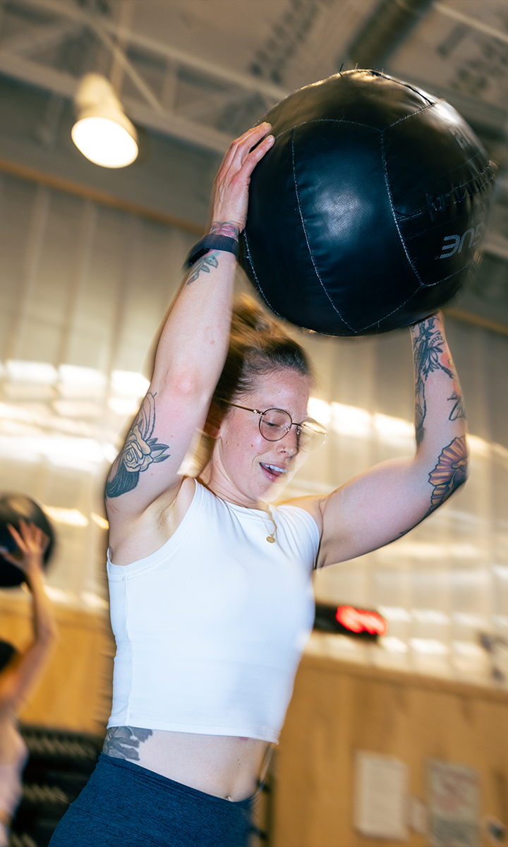 A woman with tattoos and glasses lifts a large black medicine ball above her head in the gym, showcasing strength and focus. Experience scenes like this and more with Membership Benefits at Austin Bouldering Project.