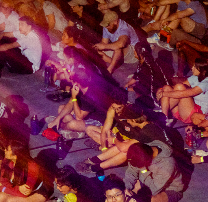A large group sits closely together on the ground at an outdoor event, illuminated by colorful, streaked lighting. Some chat while others look forward, relaxed and engaged—just like experiencing membership benefits at Austin Bouldering Project.