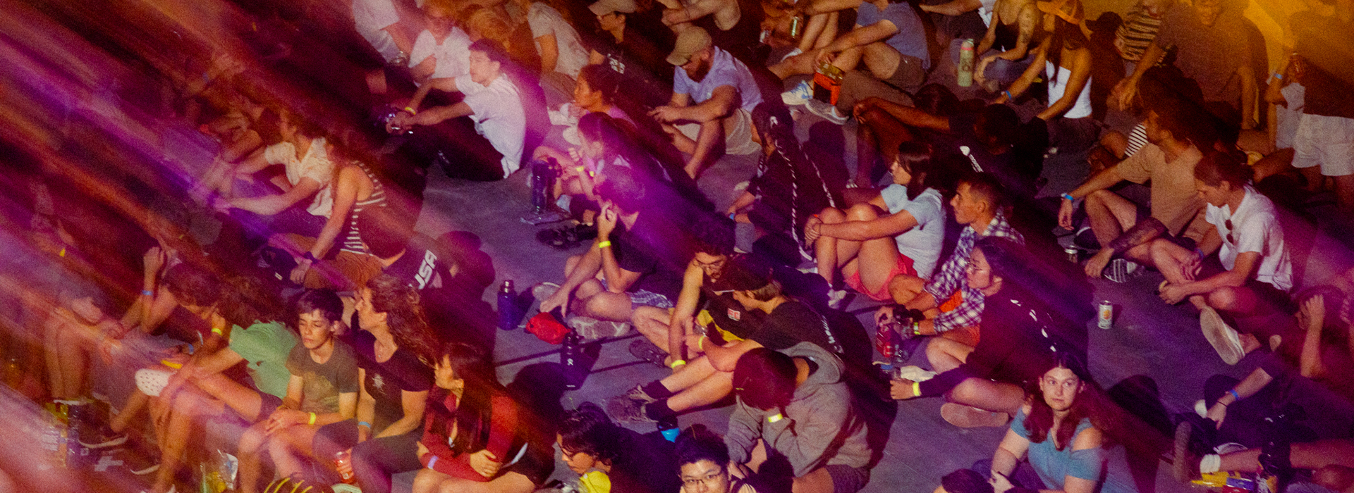 A large group sits closely together on the ground at an outdoor event, illuminated by colorful, streaked lighting. Some chat while others look forward, relaxed and engaged—just like experiencing membership benefits at Austin Bouldering Project.