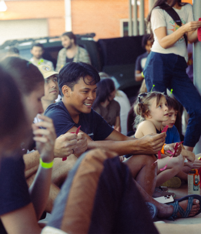 A group of people, including adults and children, sit closely together outdoors. A man in the center smiles, while a girl beside him with green hair antennas looks ahead. The scene feels casual and relaxed—much like enjoying membership benefits at Austin Bouldering Project.