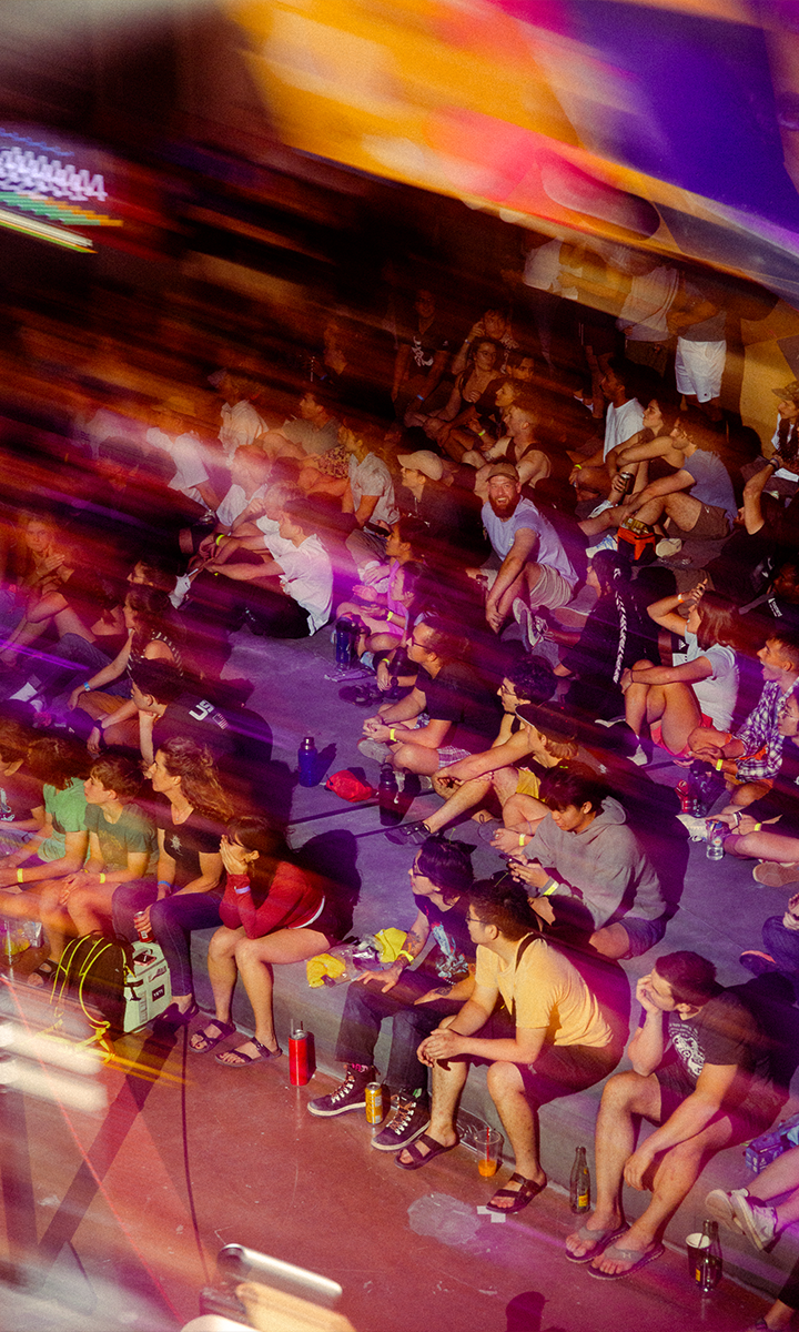 A crowd sits closely together on steps, watching an event. Colorful motion blur and streaks of light create a dynamic atmosphere. Drinks and bags are scattered around them, echoing the vibrant community spirit found in Membership Benefits at Austin Bouldering Project.