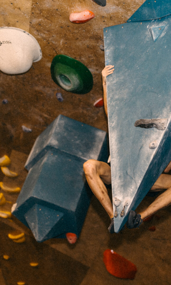 A person climbs an indoor bouldering wall, mostly hidden behind a large blue triangular hold, their chalked legs, feet, and hands gripping the holds—perhaps enjoying the perks of a discounted membership.