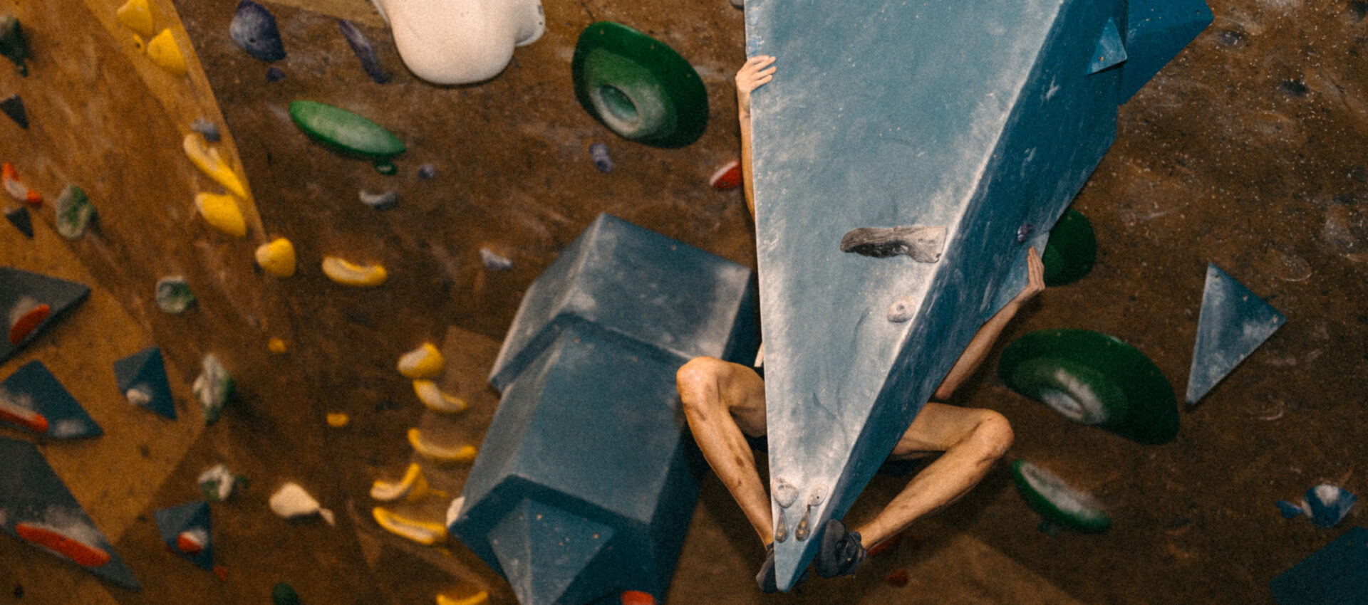 A person climbs an indoor bouldering wall, gripping a large blue triangular hold with their arms and legs. Colorful climbing holds are visible on the wall—perfect for those seeking adventure or a discounted membership.