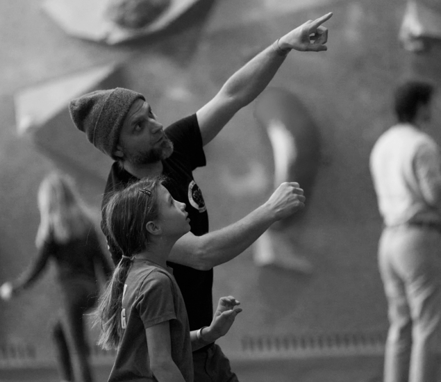 A bearded man wearing a beanie points upward while talking to a young girl, both focused, possibly discussing climbing at the Minneapolis Boulderfest competition. Two blurred people and a climbing wall are visible in the black-and-white image.