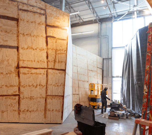 A person works inside a large industrial space with unfinished plywood walls, construction materials scattered on the floor, and sunlight streaming through tall windows. A yellow lift and a covered area are visible.