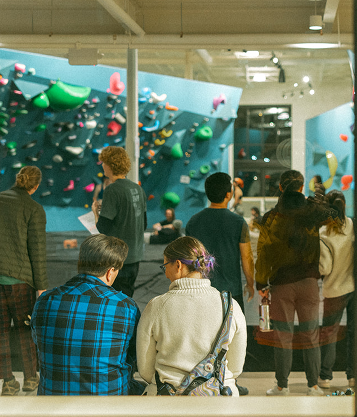 A lively, social scene unfolds at St. Paul Bouldering Project Climbing Gym, where people gather near colorful climbing walls. Two sit in the foreground, backs turned to the camera, while others stand and admire the walls.