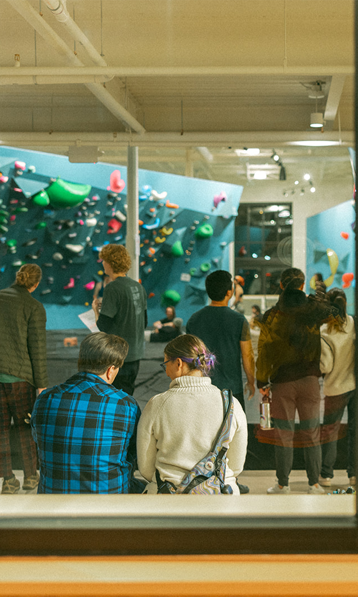 A lively, social scene unfolds at St. Paul Bouldering Project Climbing Gym, where people gather near colorful climbing walls. Two sit in the foreground, backs turned to the camera, while others stand and admire the walls.