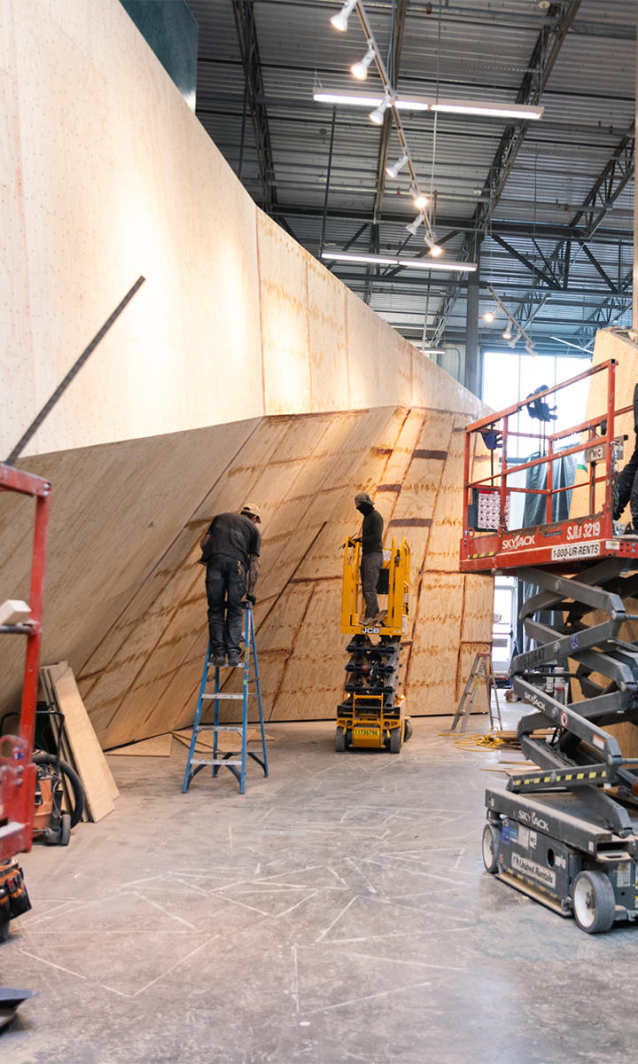 Two workers, one on a blue ladder and one on a yellow lift, install large wooden panels on a sloped wall inside a spacious warehouse with industrial lighting and scaffolding equipment nearby.