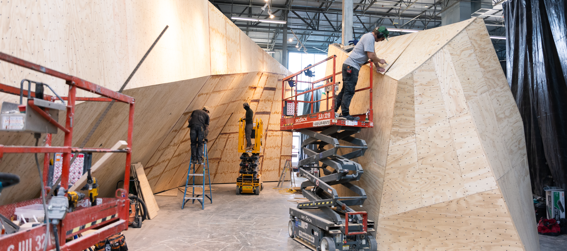 Workers use ladders and scissor lifts to construct large, angular wooden structures inside a spacious industrial building. Tools and equipment are visible on the ground.