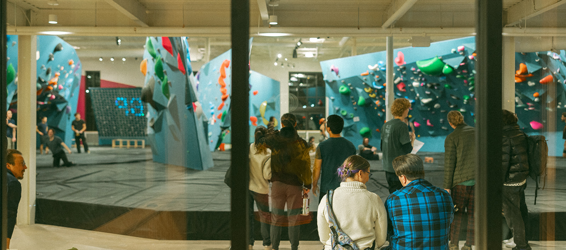 A group of people gather inside the modern St. Paul Bouldering Project Climbing Gym, standing and sitting near colorful climbing walls with holds of various shapes and sizes. The scene is viewed through large glass windows.