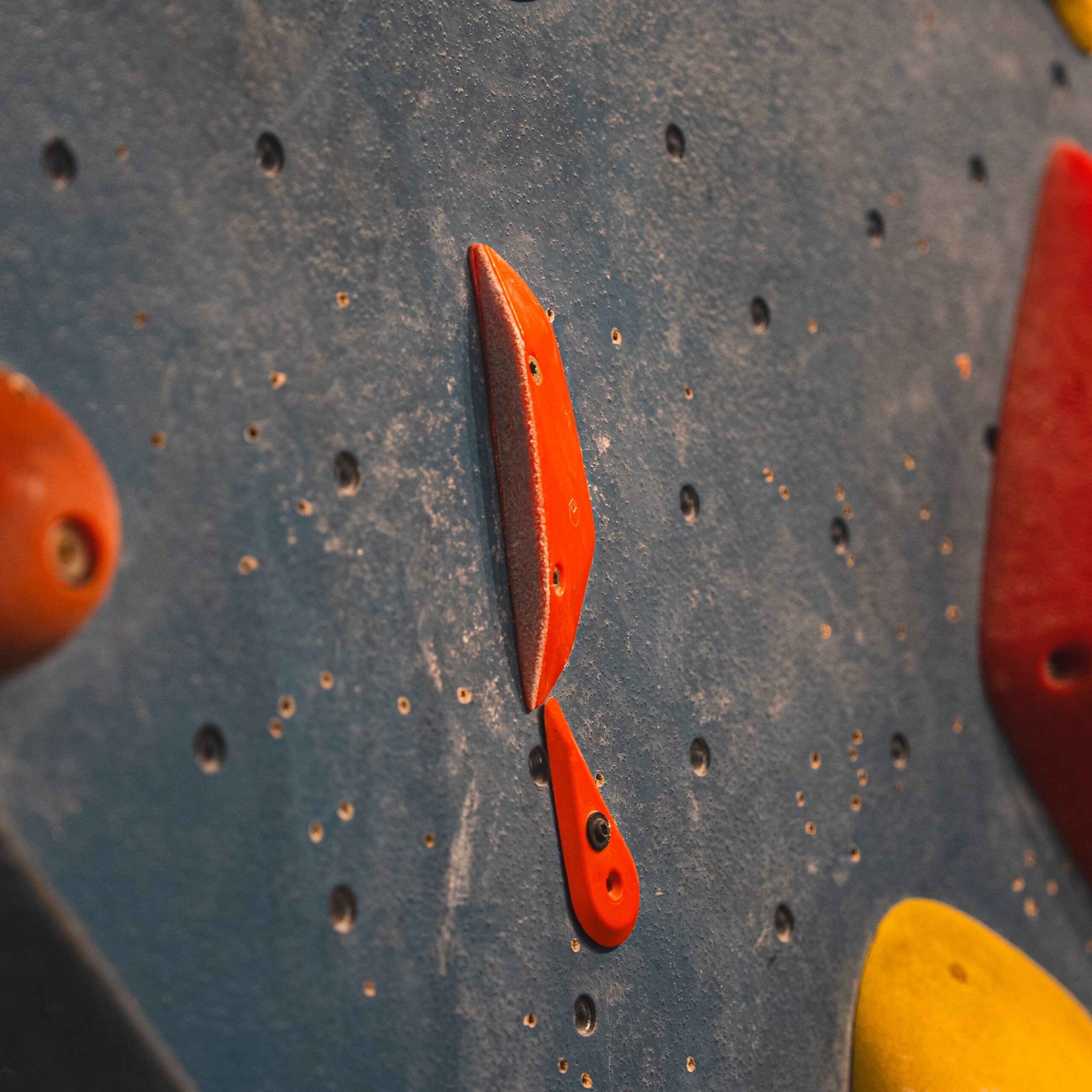 Close-up of a blue indoor climbing wall featuring rock climbing holds, including a small red handhold and a smaller red foothold, surrounded by other holds and many empty bolt holes.