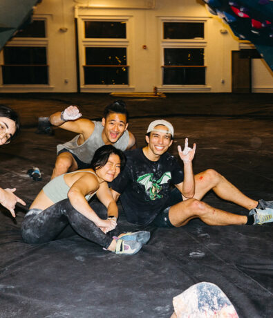 Four young adults with climbing gear and chalk on their hands pose and smile while sitting on the floor of an indoor climbing gym, enjoying Membership Benefits at Austin Bouldering Project, surrounded by colorful climbing walls and holds.