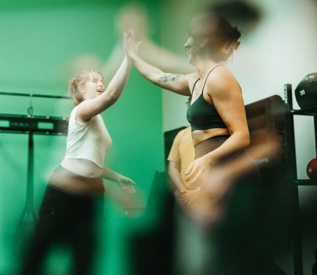 Two women in athletic wear high-five each other in a gym with green lighting, smiling and celebrating the Membership Benefits at Austin Bouldering Project. The image is softly blurred, adding dynamic energy with fitness equipment in the background.
