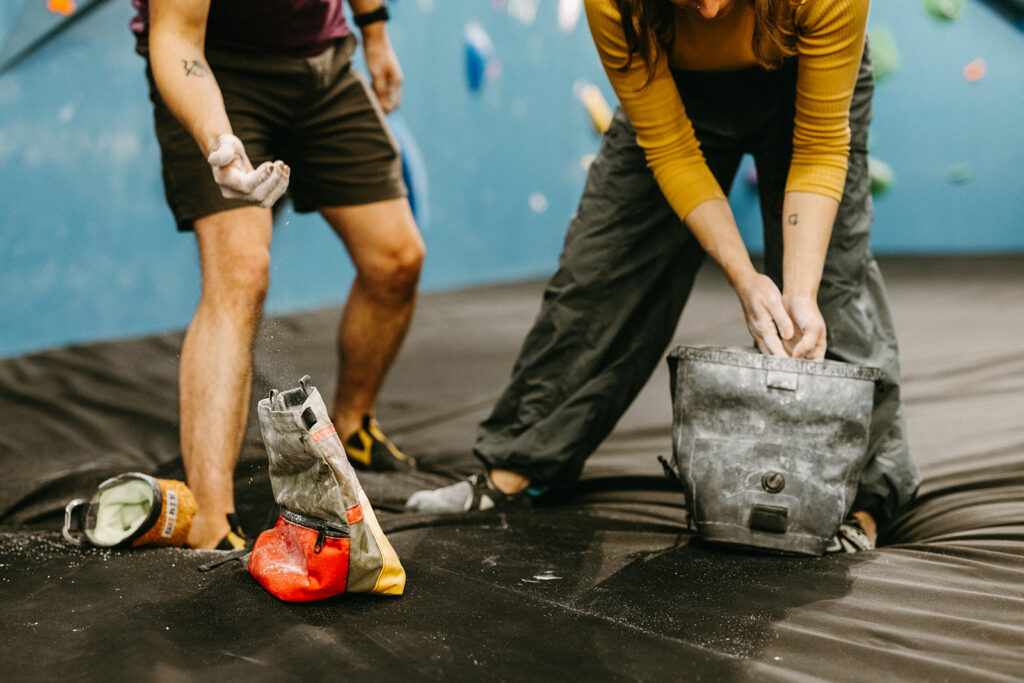 Two people stand on a padded floor at an indoor climbing gym, applying chalk to their hands as they discuss climbing grades. Chalk bags and climbing shoes are visible, with colorful holds on the blue wall in the background.