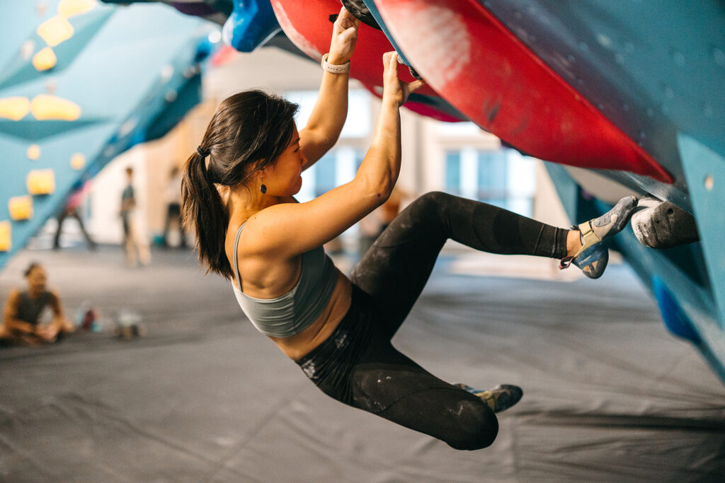 A woman wearing athletic clothing and climbing shoes is bouldering on an indoor climbing wall, gripping red holds with her hands and feet on blue footholds, as she tackles different climbing grades. The background shows padded flooring and other people.