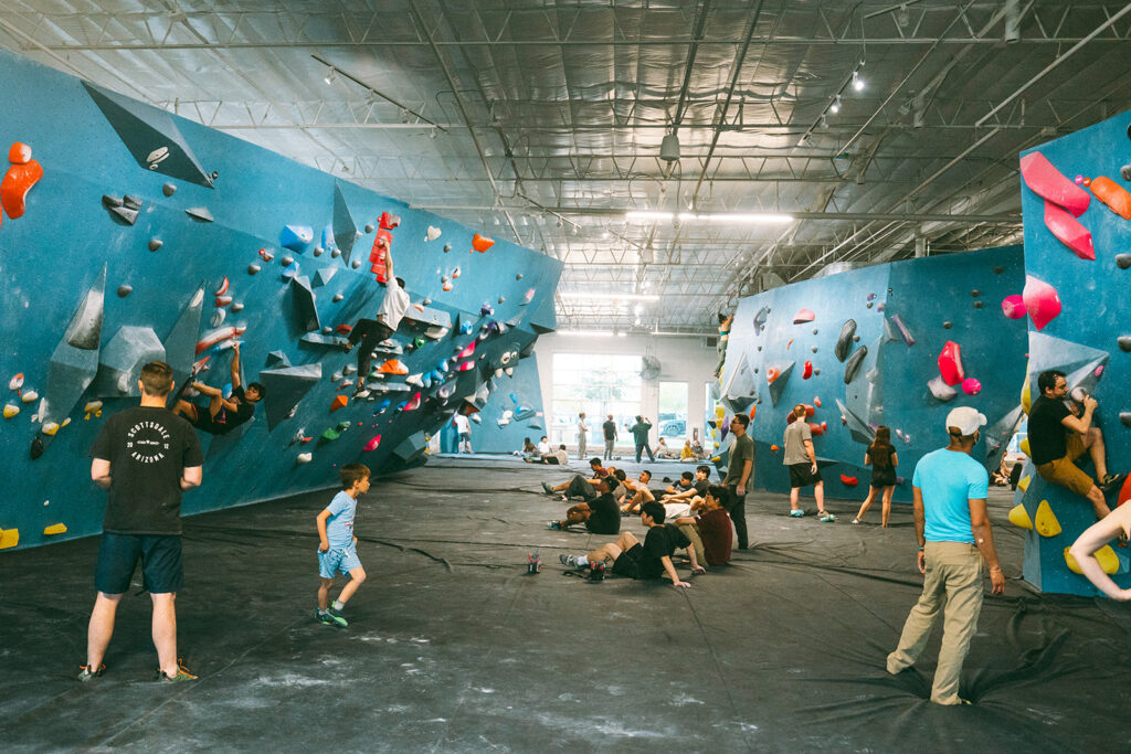 Indoor climbing gym with people of various ages bouldering on blue walls with colorful rock climbing holds. Some are climbing, while others sit or stand on padded floors. Children are also present, and natural light streams through large windows.