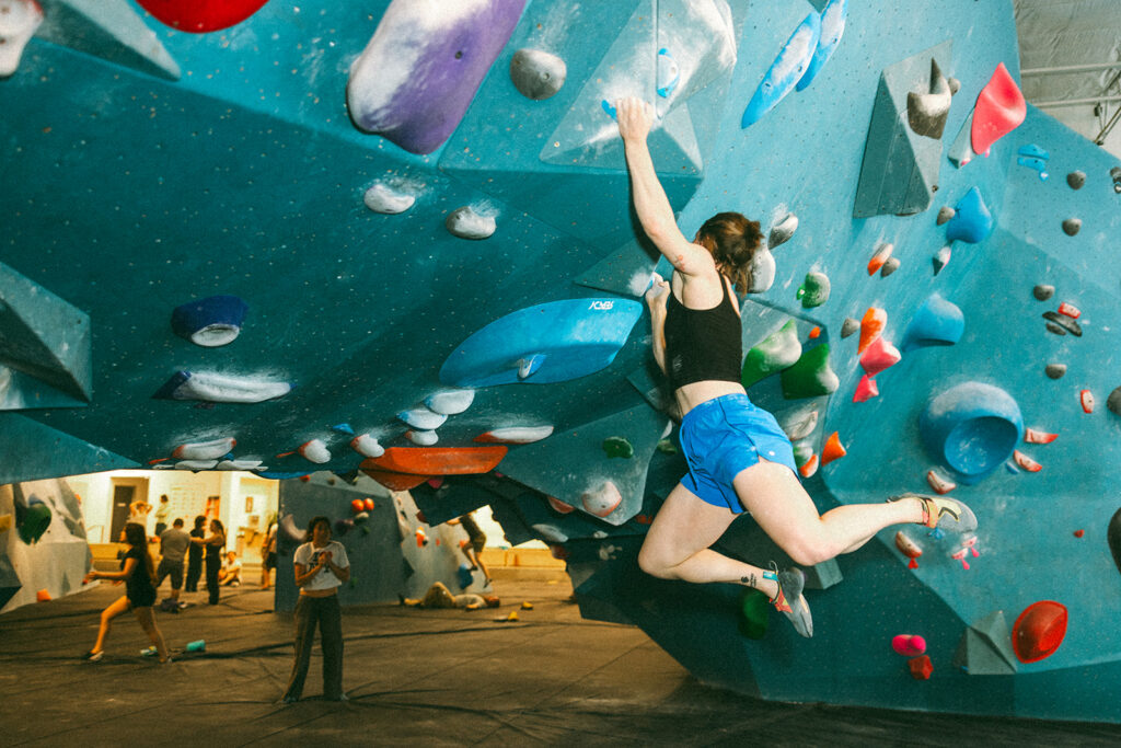 A person in blue shorts and a black tank top climbs an overhanging indoor bouldering wall, gripping vibrant rock climbing holds with extended arms while others watch and climb in the background.