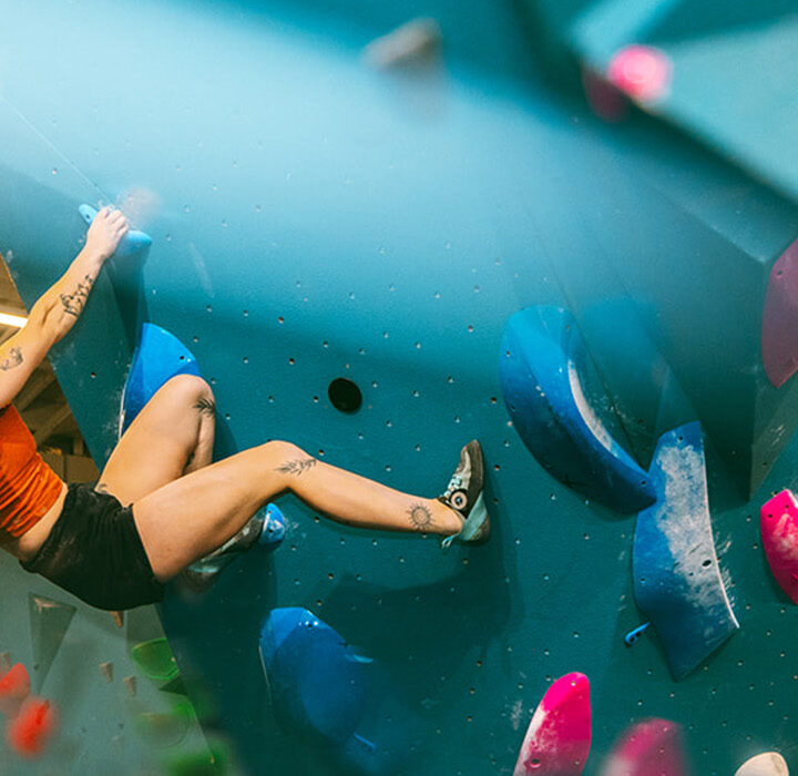 A person with tattoos climbs an indoor bouldering wall, gripping colorful holds. They wear a red tank top, black shorts, and climbing shoes, their hair loose in a brightly lit gym—perfect for making the most of a summer membership.